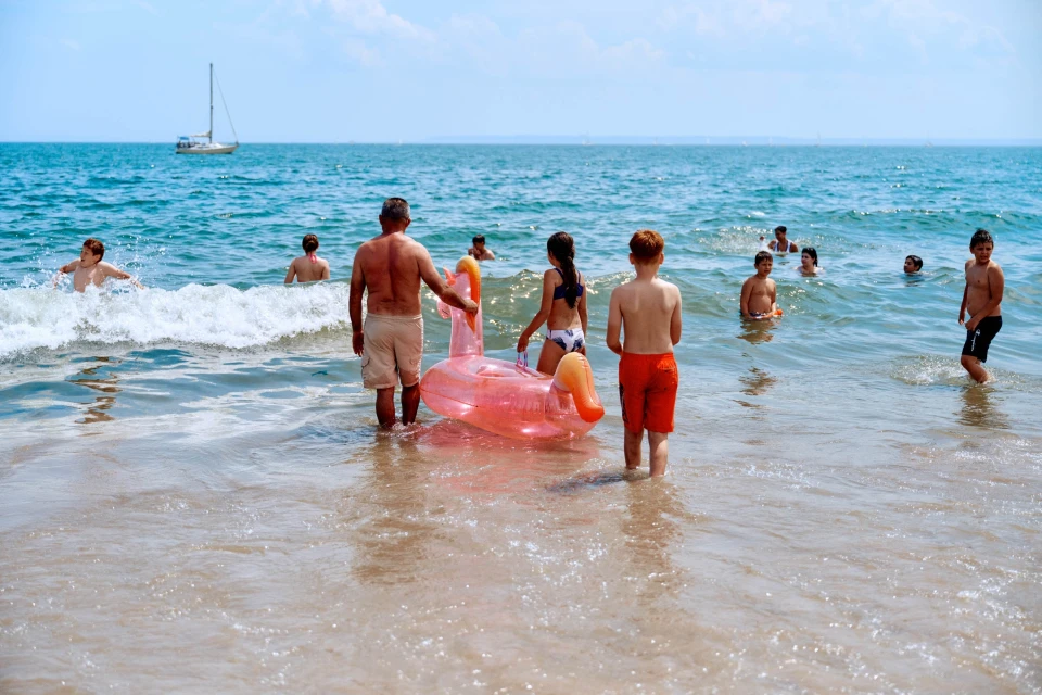 Several people, including children and adults, are enjoying a sunny day at the beach, some wading in the water with a pink flamingo float.