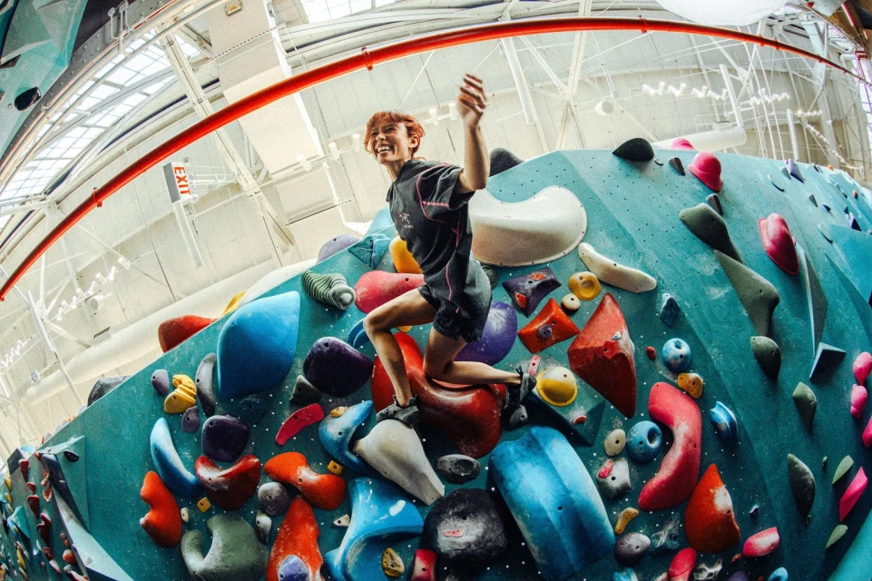 Young adult climbs a blue indoor rock wall with colorful holds in a fitness gym with white ceiling, using dynamic movement and upper body strength.