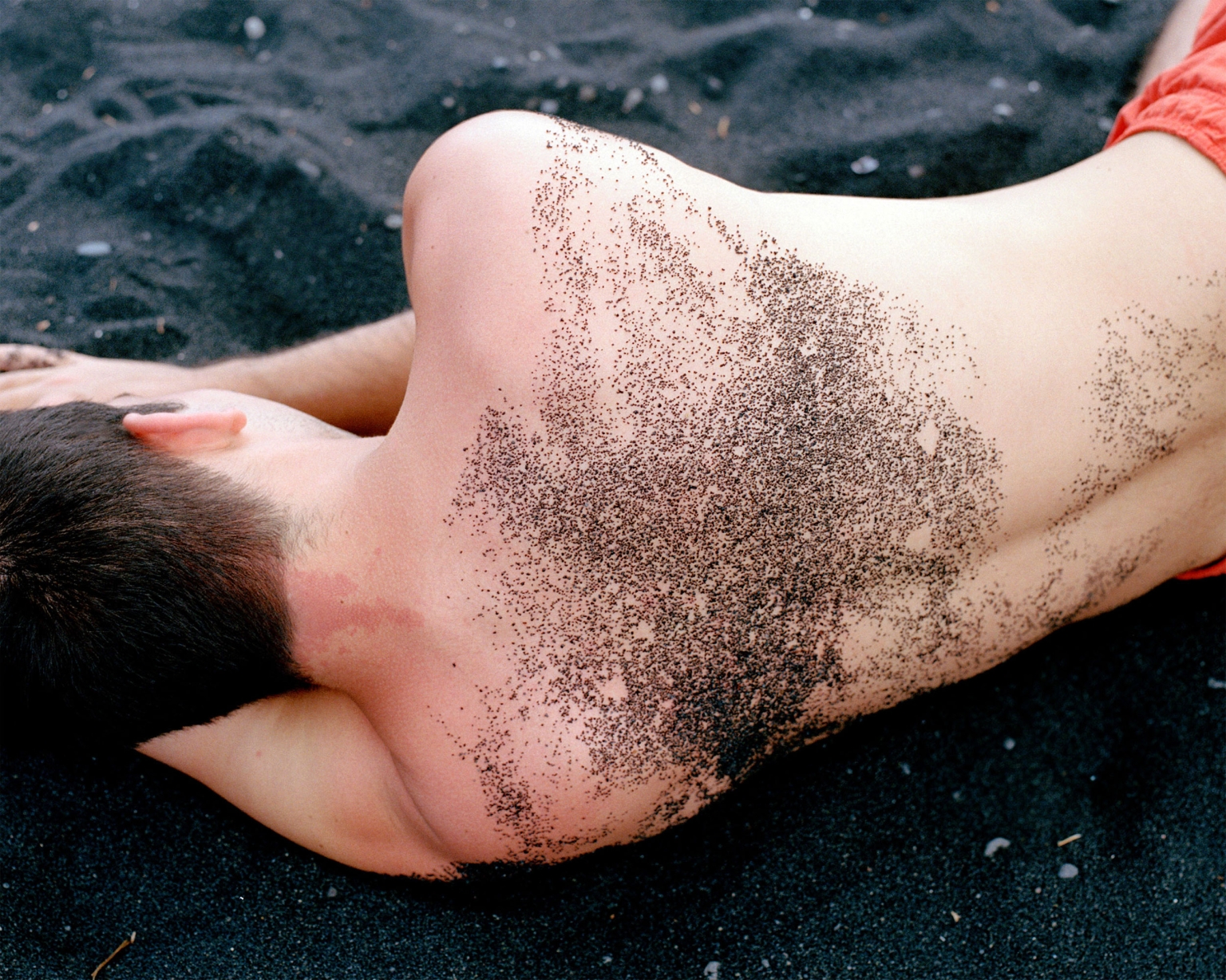 Man lies on his side on a black sand beach, with sand stuck to his skin; red shorts are visible. The image is a close-up shot.