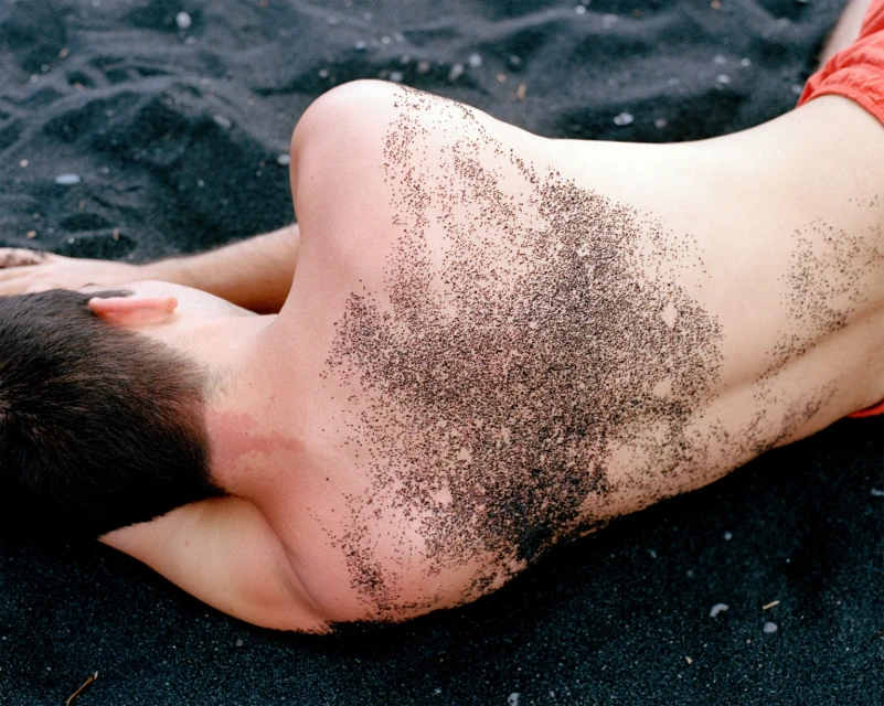 Man lies on his side on a black sand beach, with sand stuck to his skin; red shorts are visible. The image is a close-up shot.