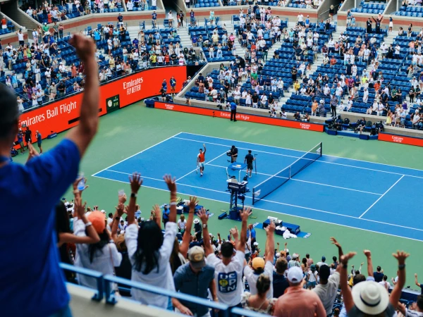 Tennis player wins match at crowded stadium with cheering fans, blue court, and orange Emirates advertising banner in the background.