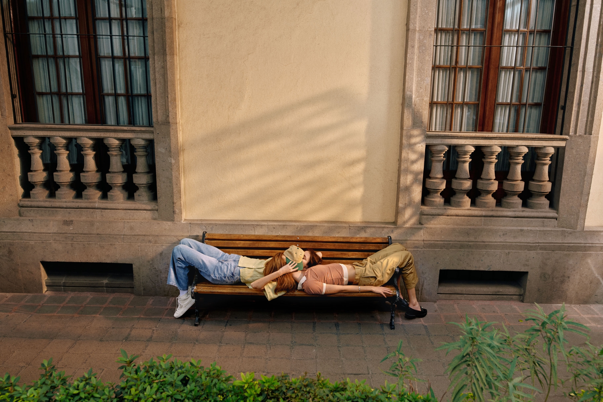 Two young women lie head-to-head on a bench in front of a pale building with windows, their bodies stretched out, enjoying the sunny afternoon.