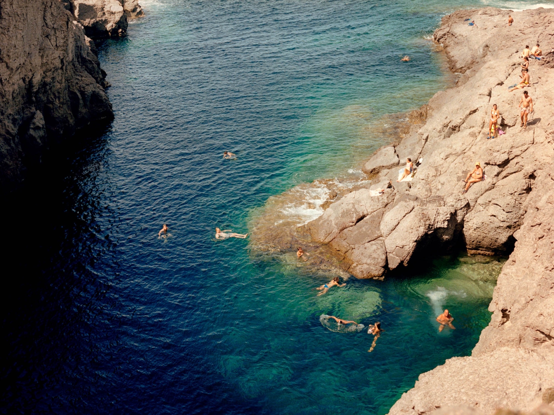 Aerial view of a rocky coastline with people swimming and sunbathing in clear turquoise water on a sunny day, creating a summer scene.