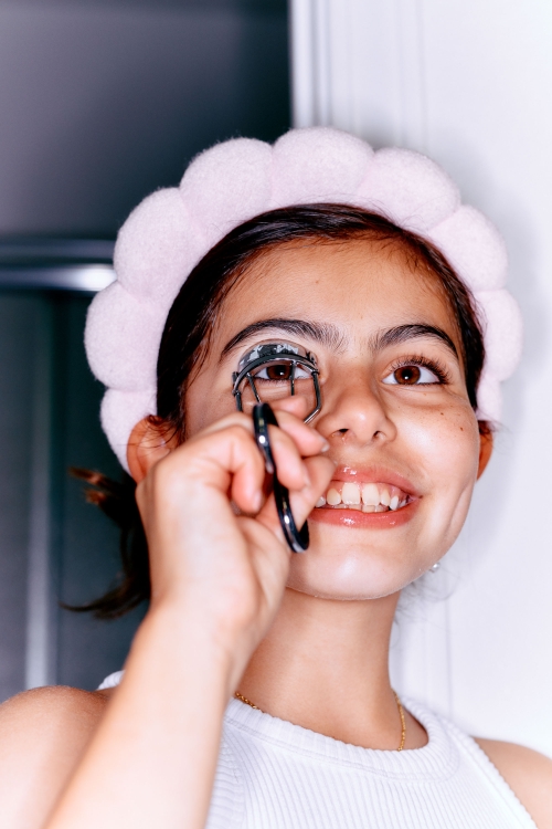 A teenage girl with a pink headband is shown in a medium shot, curling her eyelashes with a black eyelash curler in an indoor setting.