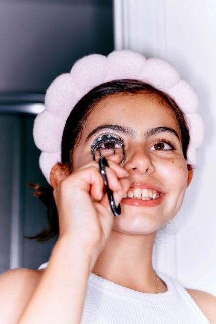 A teenage girl with a pink headband is shown in a medium shot, curling her eyelashes with a black eyelash curler in an indoor setting.