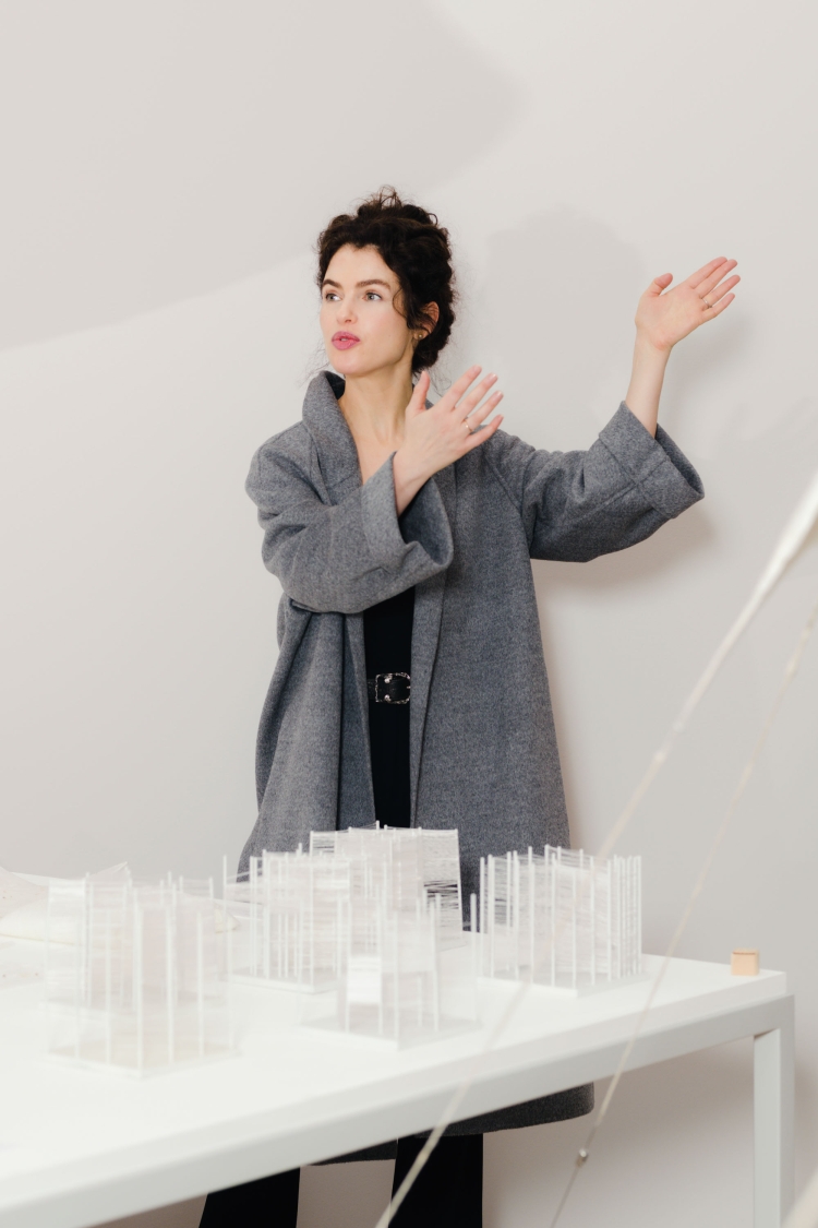 Adult woman in a grey coat stands near a scale model of a building on a table indoors, with a white wall in the background.