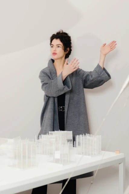 Adult woman in a grey coat stands near a scale model of a building on a table indoors, with a white wall in the background.