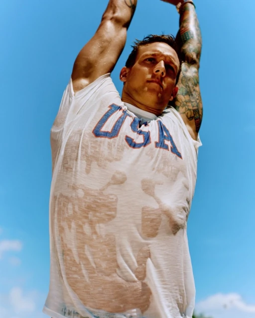 Low-angle portrait of muscular adult male with USA shirt stretching arms overhead in sunny outdoor setting with blue sky background.