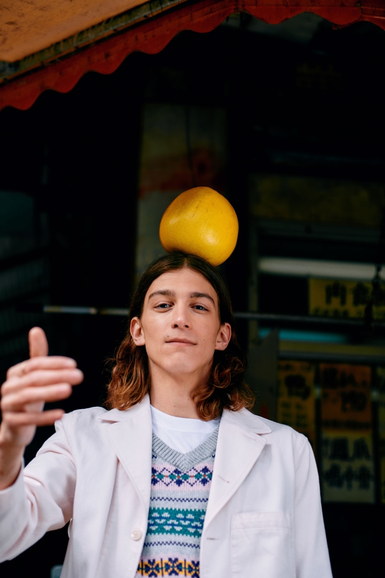 Young adult balances yellow fruit on head wearing a patterned sweater and lab coat in front of a dark urban background.