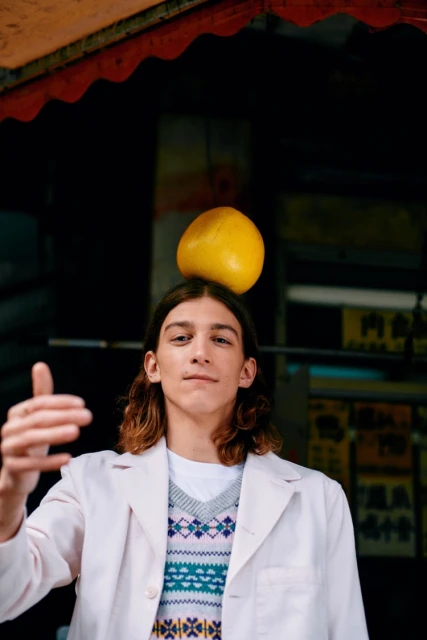 Young adult balances yellow fruit on head wearing a patterned sweater and lab coat in front of a dark urban background.