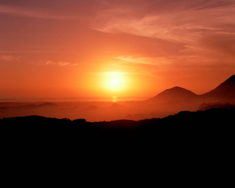 Vibrant sunset over silhouetted mountains and dark hills with an orange sky and a hazy horizon; a scenic view.