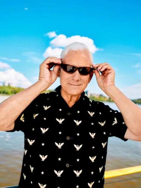 Older man in patterned short-sleeved shirt adjusts sunglasses against a backdrop of blue skies and river, creating a summery portrait.