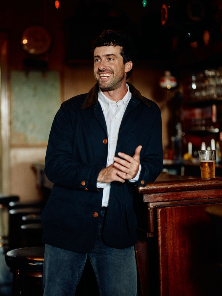 Man with a beard and dark hair in a navy blazer and jeans leans against a wooden bar indoors, smiling, with a beer in the background.