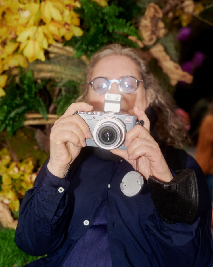 A person with eyeglasses holds a gray camera in front of a display of yellow orchids and greenery.