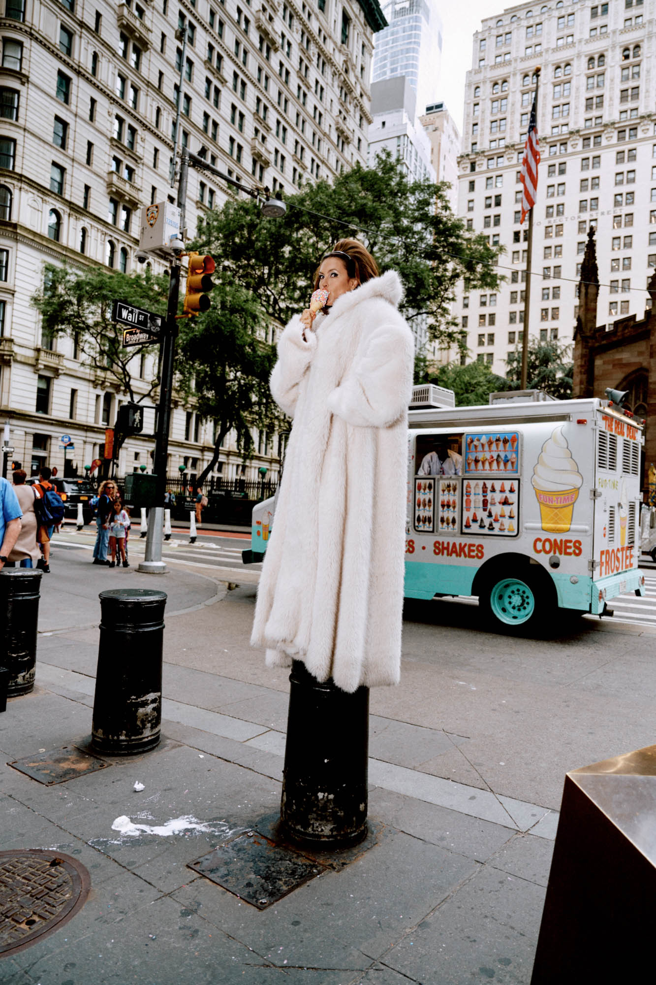 Woman in a fur coat poses on a bollard on a city street near an ice cream truck and tall buildings in an urban environment.