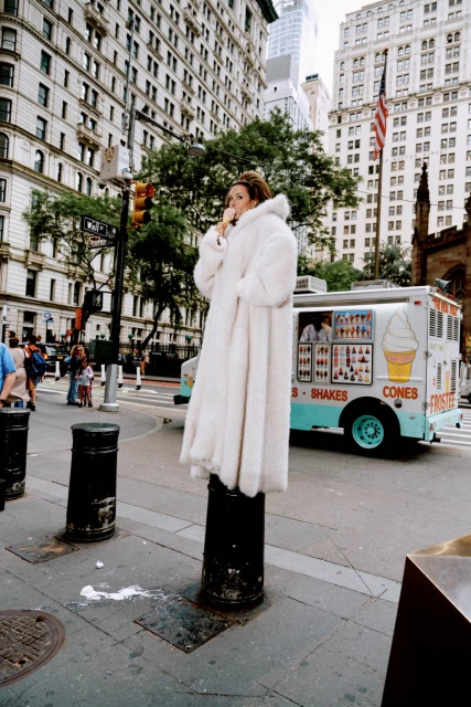 Woman in a fur coat poses on a bollard on a city street near an ice cream truck and tall buildings in an urban environment.