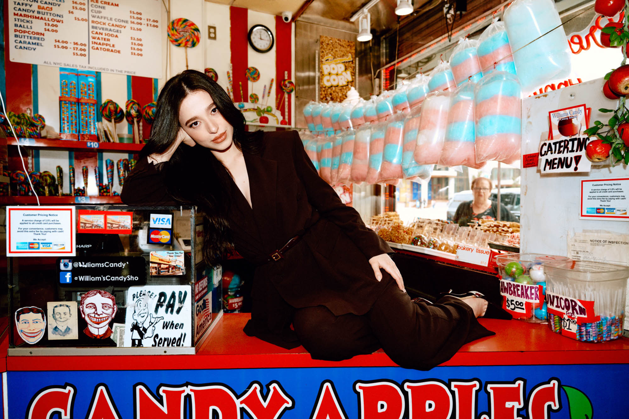 Adult female in a brown suit poses in front of a candy shop with candy apples, cotton candy, and various candies on display.