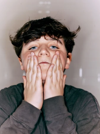 Boy with brown hair and gray shirt touching his face with both hands in a portrait with a neutral background and soft light.