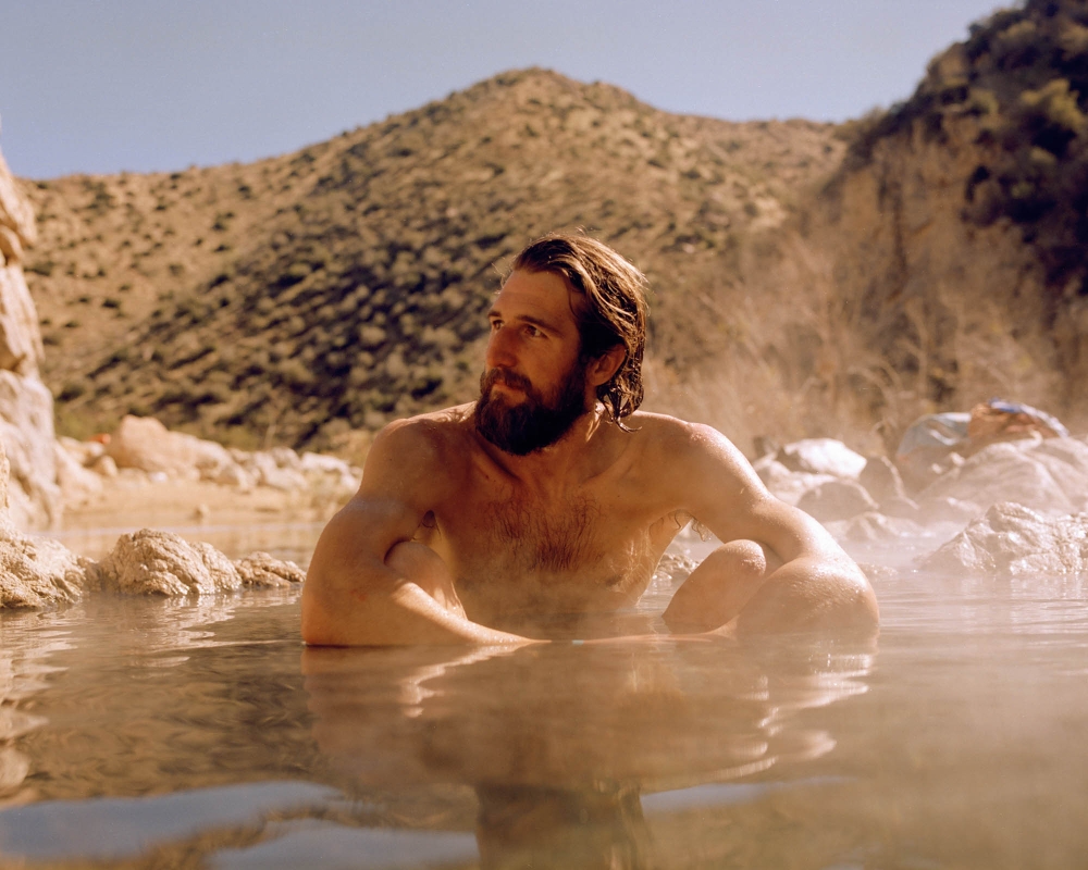 Man with a beard relaxing in steaming water in a natural outdoor hot spring with mountains under a clear blue sky in the background.
