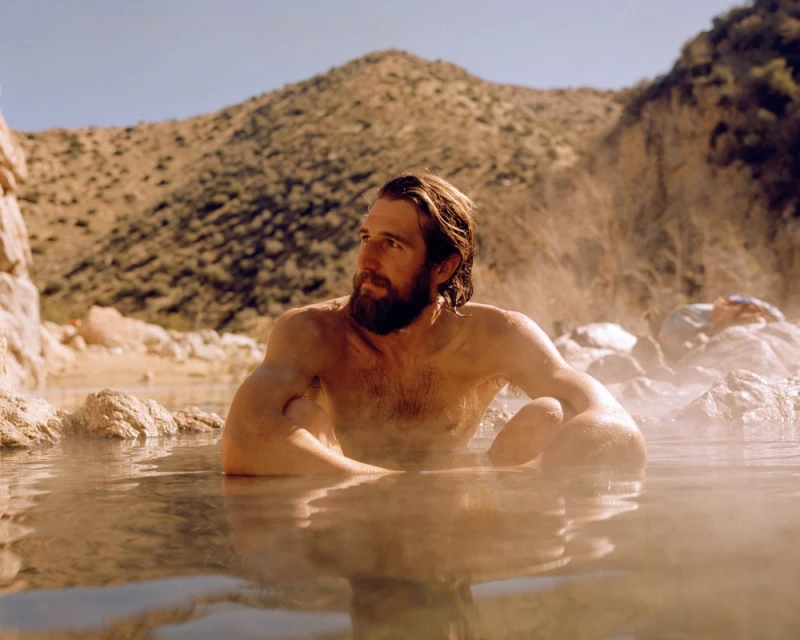Man with a beard relaxing in steaming water in a natural outdoor hot spring with mountains under a clear blue sky in the background.