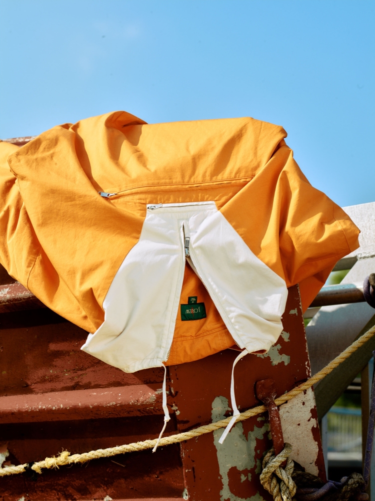 Close-up of an open orange jacket with white lining, showing a small green patch logo, against a background of blue sky and rusted metal.