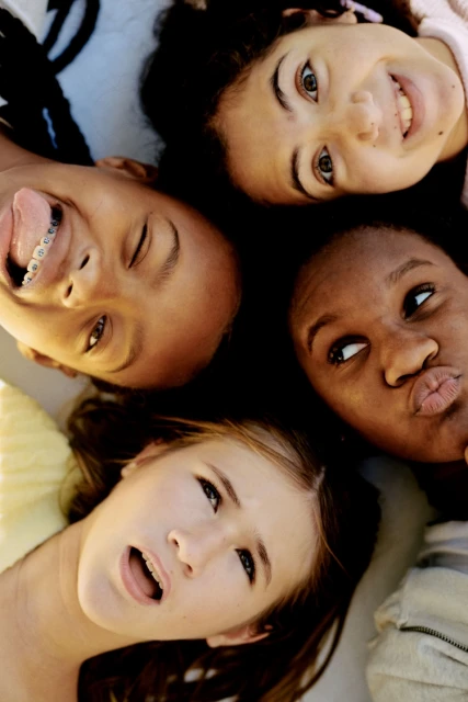 Four smiling diverse teenage girls lie in a circle with their heads together, displaying various facial expressions in an eye-level shot.