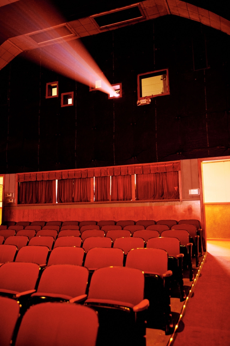 Rows of unoccupied red seats in a dark theatre, with light beam from projector, casting a reddish hue, creating a dramatic and quiet scene.