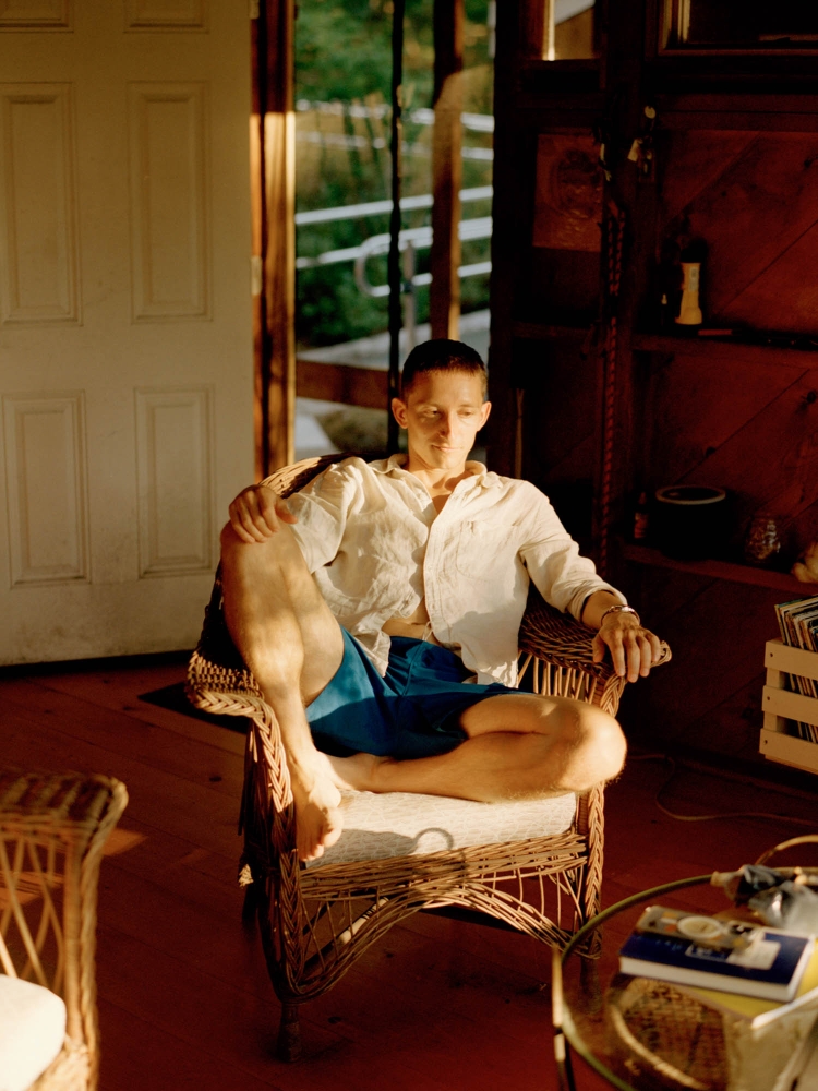 Man seated in a wicker chair in an indoor setting, wearing a white shirt and blue shorts, illuminated by natural light.