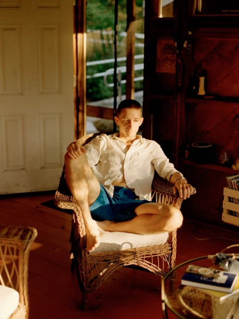 Man seated in a wicker chair in an indoor setting, wearing a white shirt and blue shorts, illuminated by natural light.