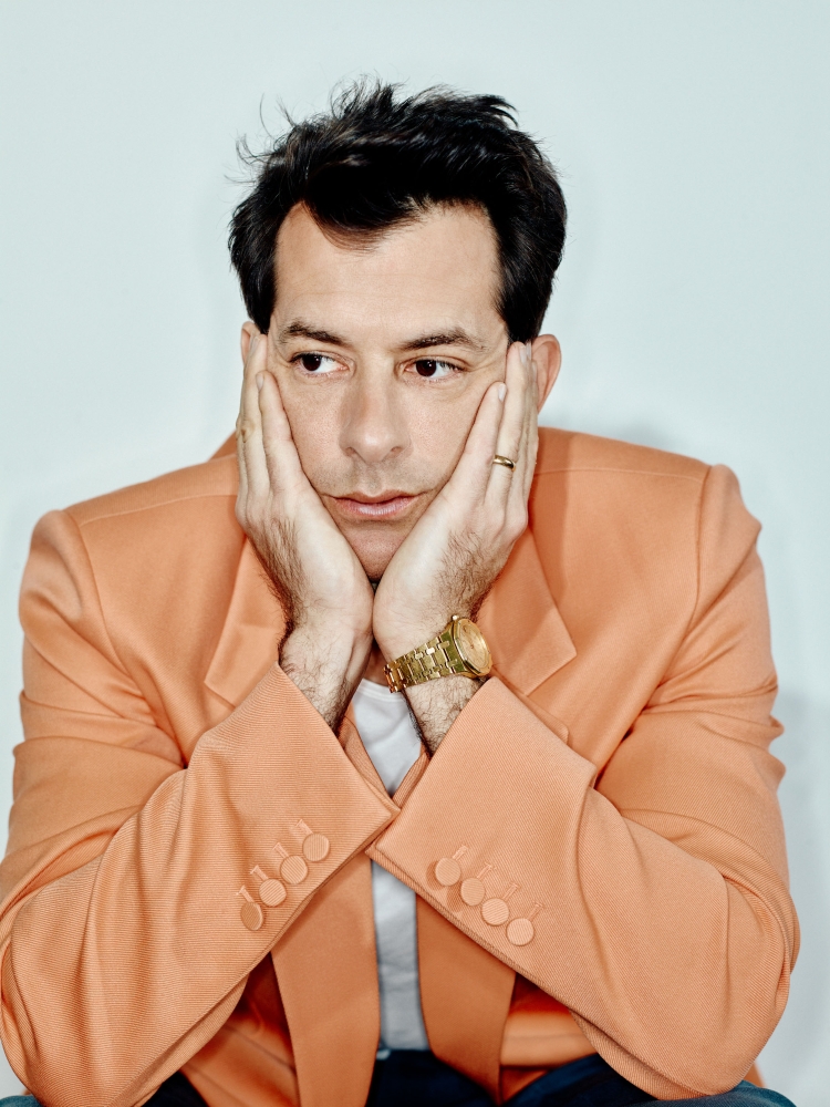 Portrait of adult male with dark hair in peach blazer with hands on his face and light expression in a photo studio.