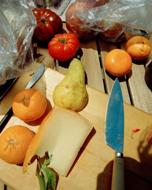 Overhead shot of a wooden table featuring a cheese wedge, pear, tomatoes, and oranges, complemented by knives and plastic produce bags.