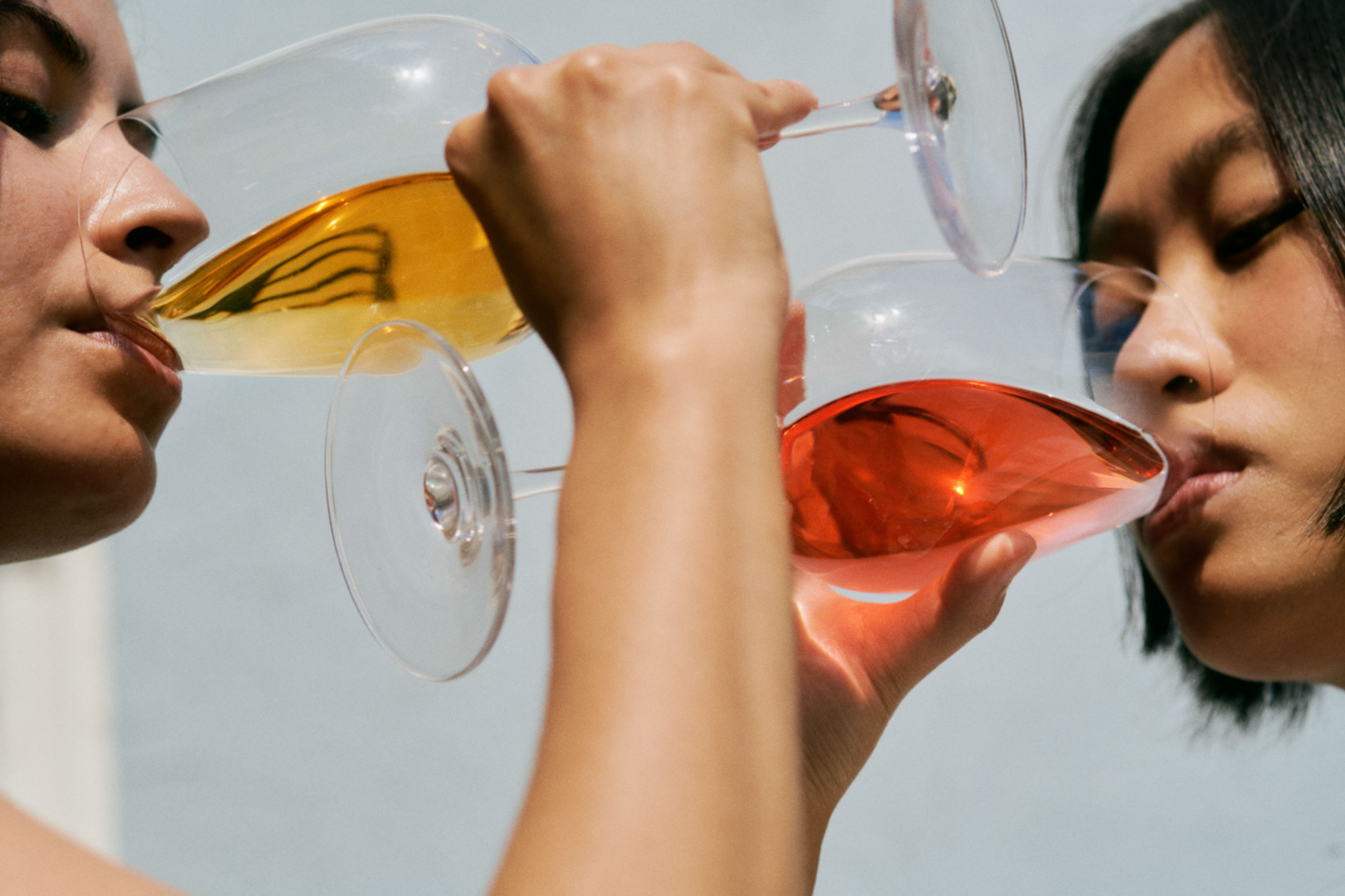 Two women are indoors drinking colored liquid from clear glasses in close-up, with a neutral background providing soft lighting.
