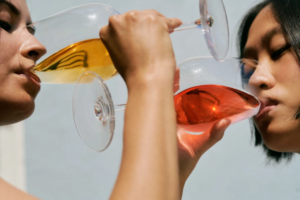 Two women are indoors drinking colored liquid from clear glasses in close-up, with a neutral background providing soft lighting.