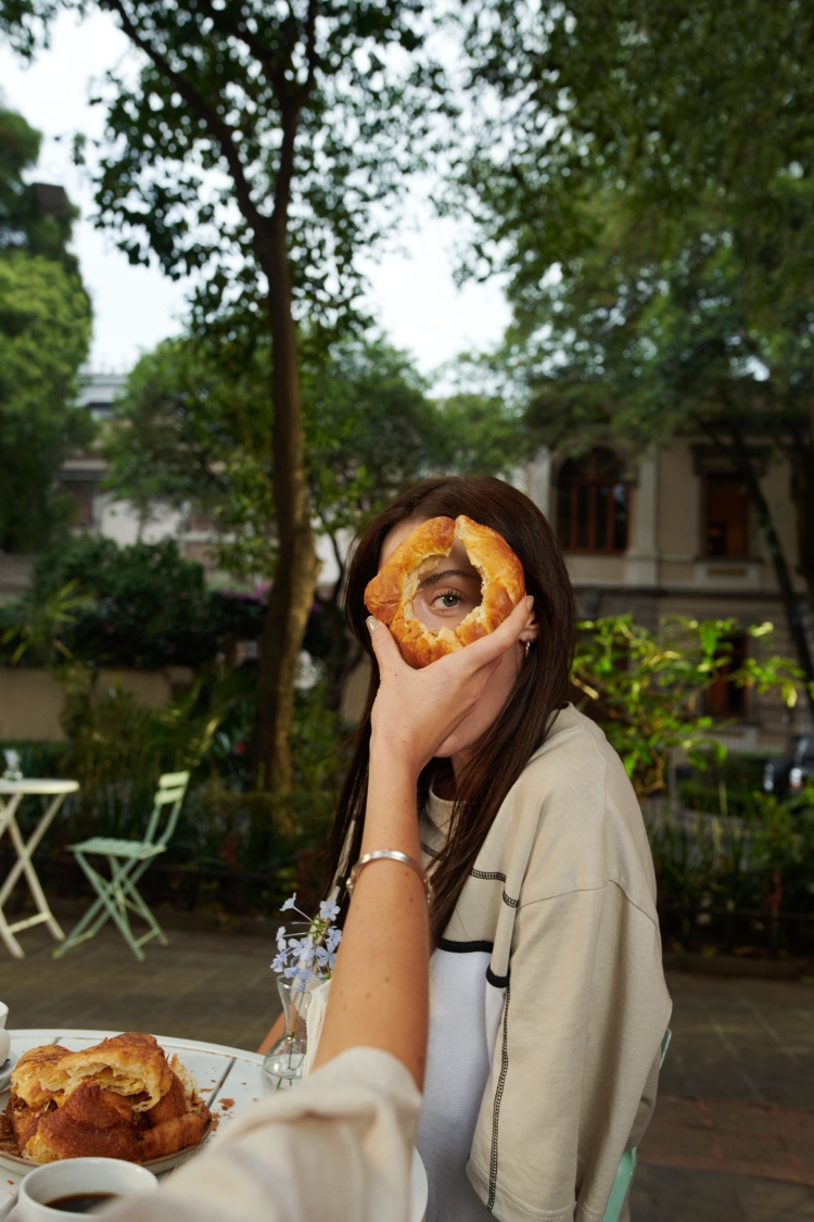 A woman is holding a round pastry in front of her face, obscuring one eye, while sitting at a table with pastries outdoors.