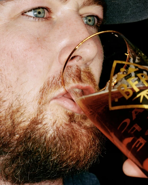 Close-up of an adult male drinking from a glass of amber beer, showcasing his red beard, and clear blue eyes, against a dark background.