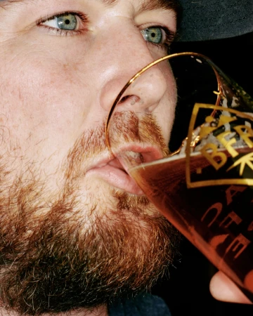 Close-up of an adult male drinking from a glass of amber beer, showcasing his red beard, and clear blue eyes, against a dark background.