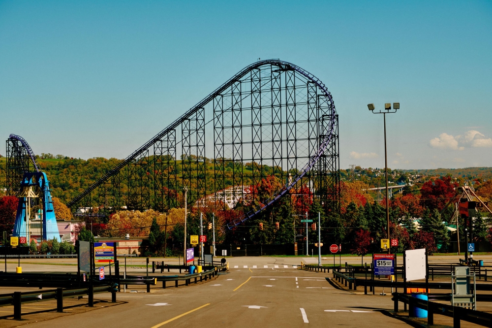 Wide shot of a black roller coaster curving through a park with trees in autumn, seen from an empty asphalt parking lot on a sunny day.