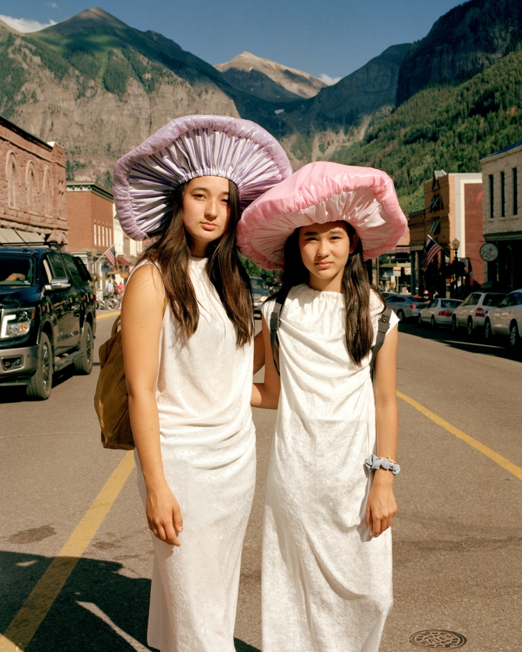 Two young women wearing white dresses and large mushroom-shaped hats standing on a sunny street with mountains in the background