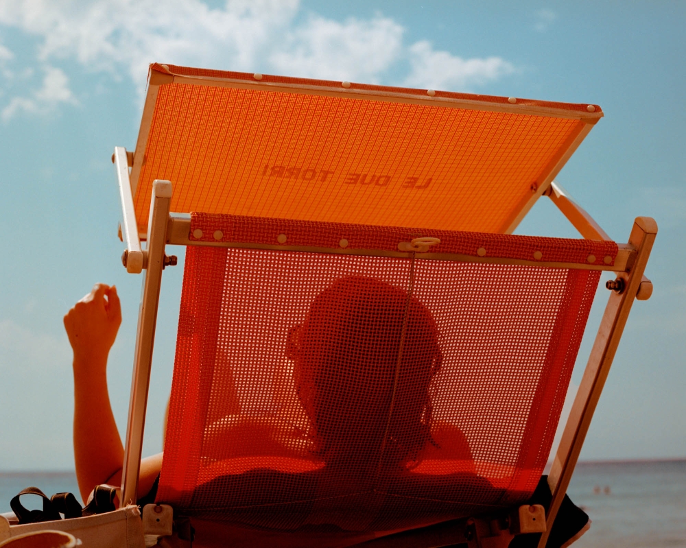 Person relaxes in an orange mesh beach chair facing the ocean under a sunny sky.  The chair has a sunshade and wooden frame.