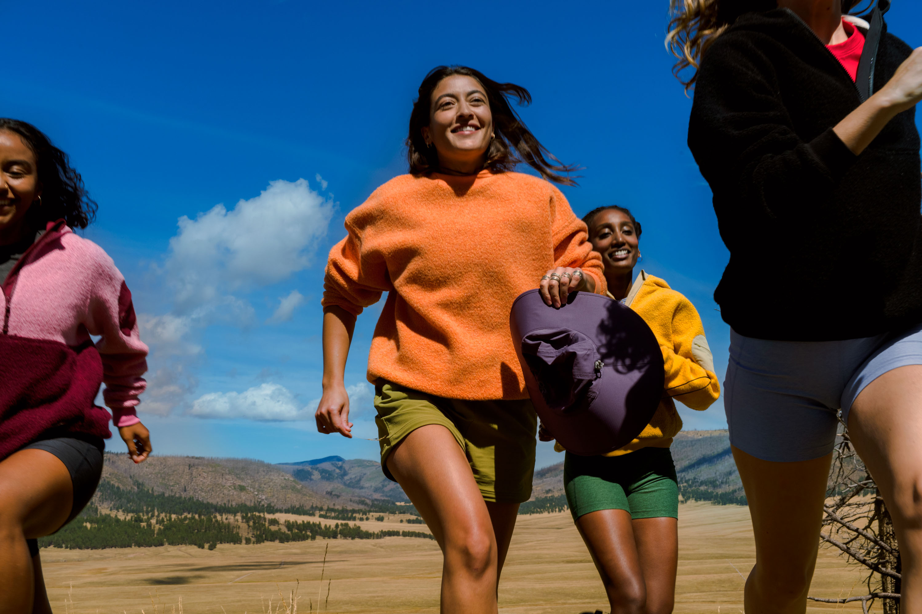 A group of four smiling women are running toward the viewer on a grassy hill under a sunny blue sky in an outdoor setting.