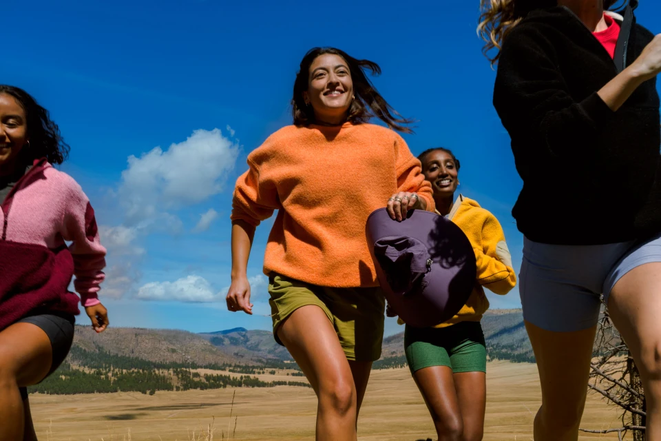 A group of four smiling women are running toward the viewer on a grassy hill under a sunny blue sky in an outdoor setting. 