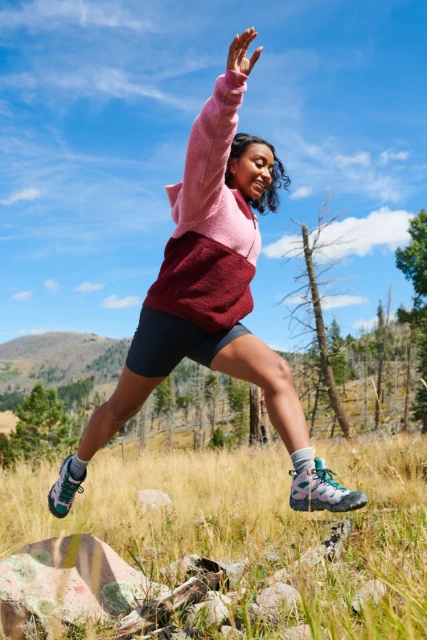 Athletic woman mid-air jumping in a mountain meadow with hiking boots, shorts, and color-blocked hooded fleece jacket against a blue sky.