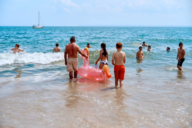 Sunny beach scene with multiple people wading and swimming in the ocean, featuring an inflatable pink flamingo toy and a distant sailboat.