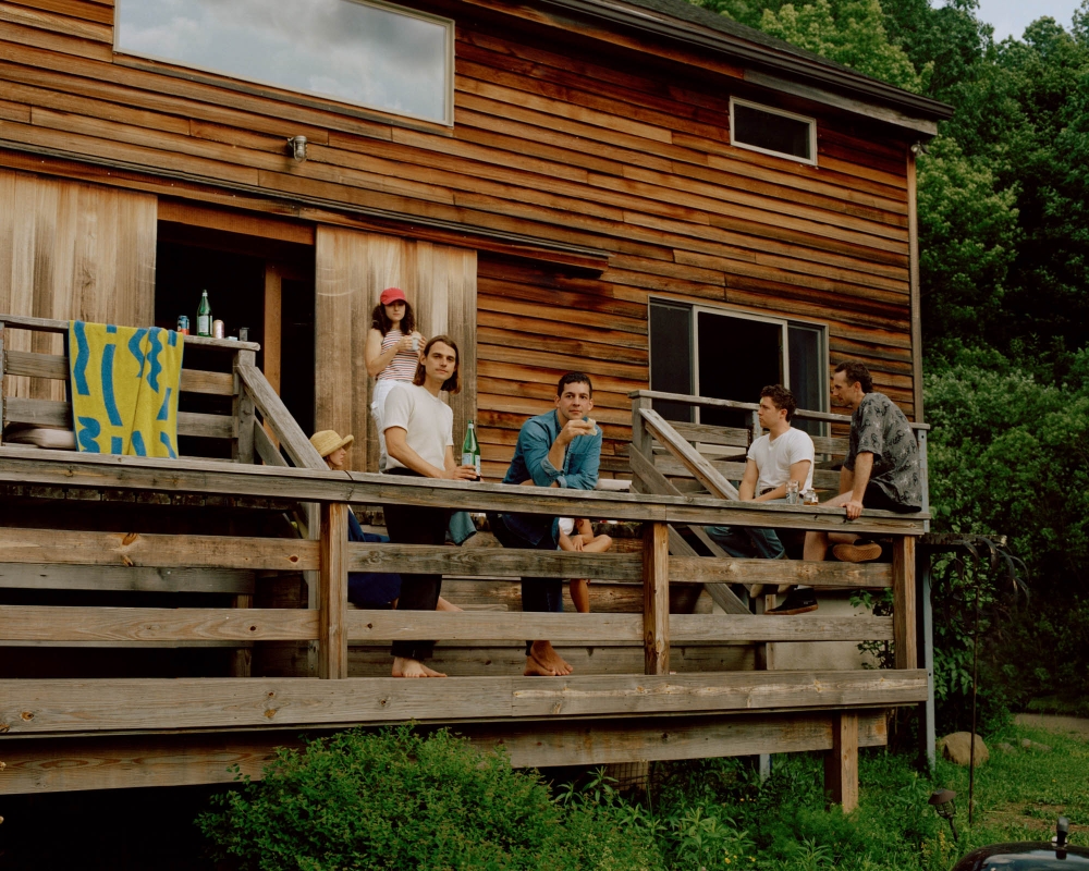 Five adults relaxing on a wood porch of a two-story wood house on a bright day with surrounding trees.
