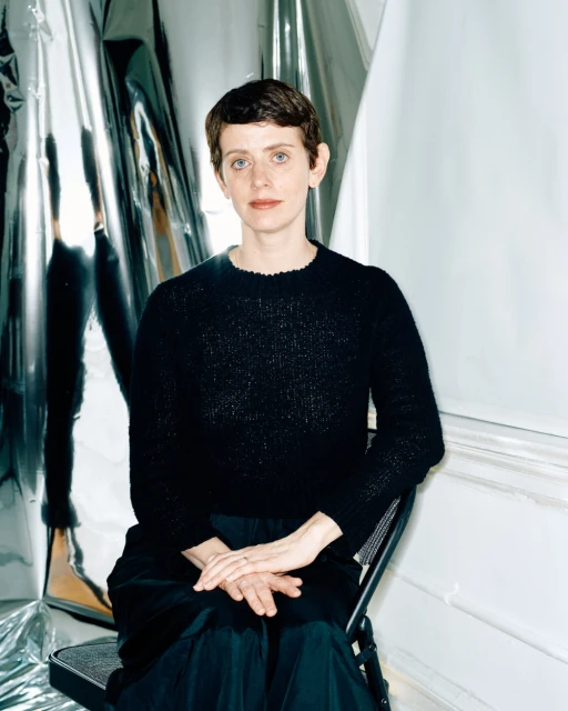 Indoor portrait of an adult woman with short hair, wearing a dark sweater, sitting in a chair in front of a reflective backdrop.