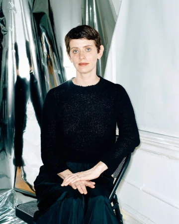 Indoor portrait of an adult woman with short hair, wearing a dark sweater, sitting in a chair in front of a reflective backdrop.