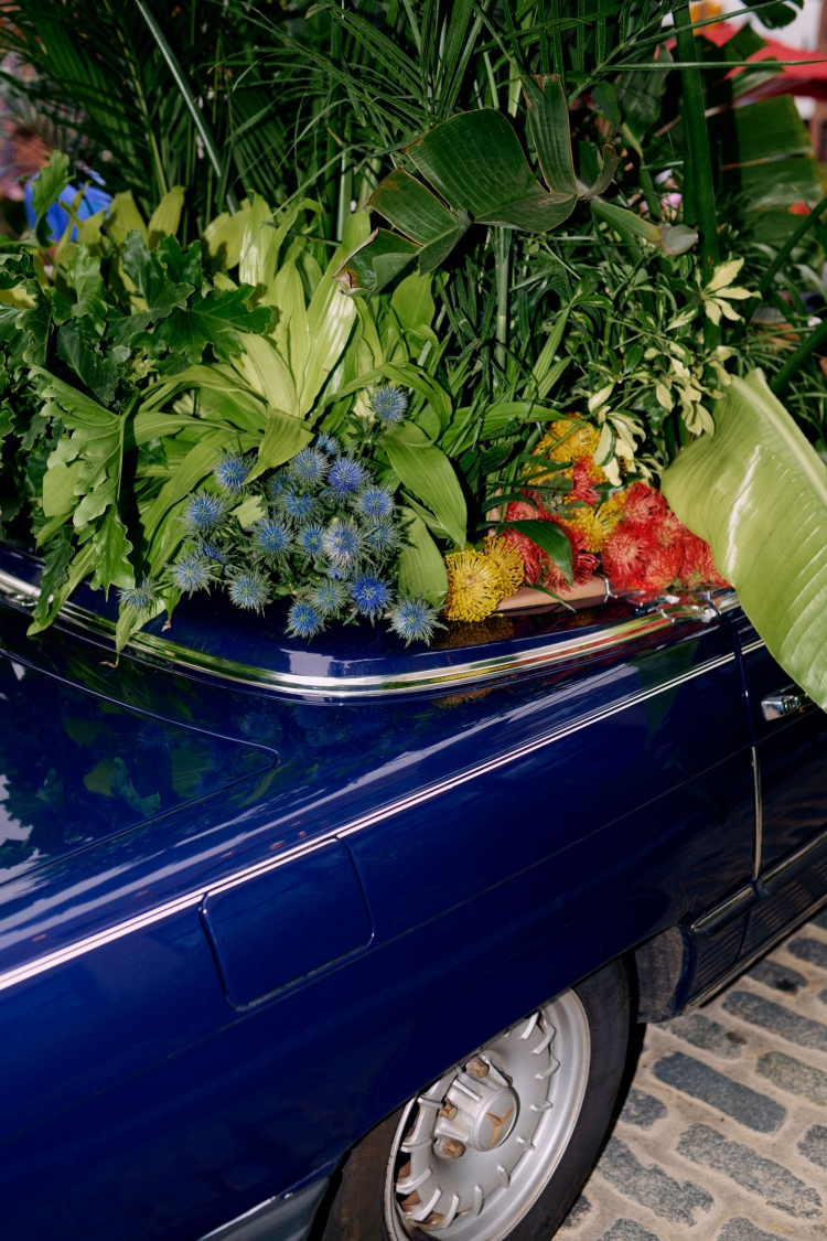 Botanical display featuring a variety of green foliage and colorful flowers arranged in the trunk of a dark blue classic car.