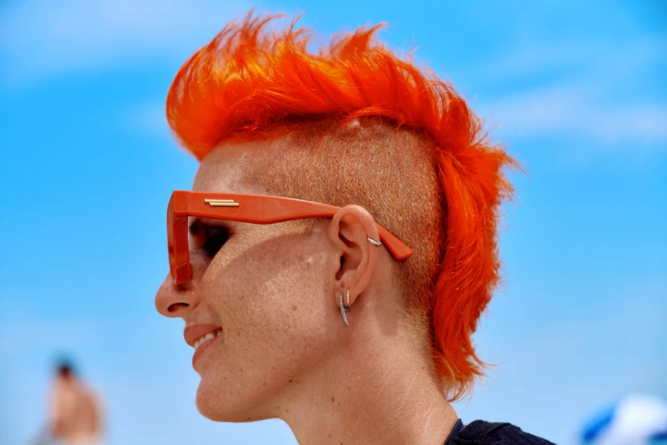 Woman with orange mohawk and sunglasses poses outdoors against a bright blue sky on a sunny day, showcasing a bold and vibrant style.