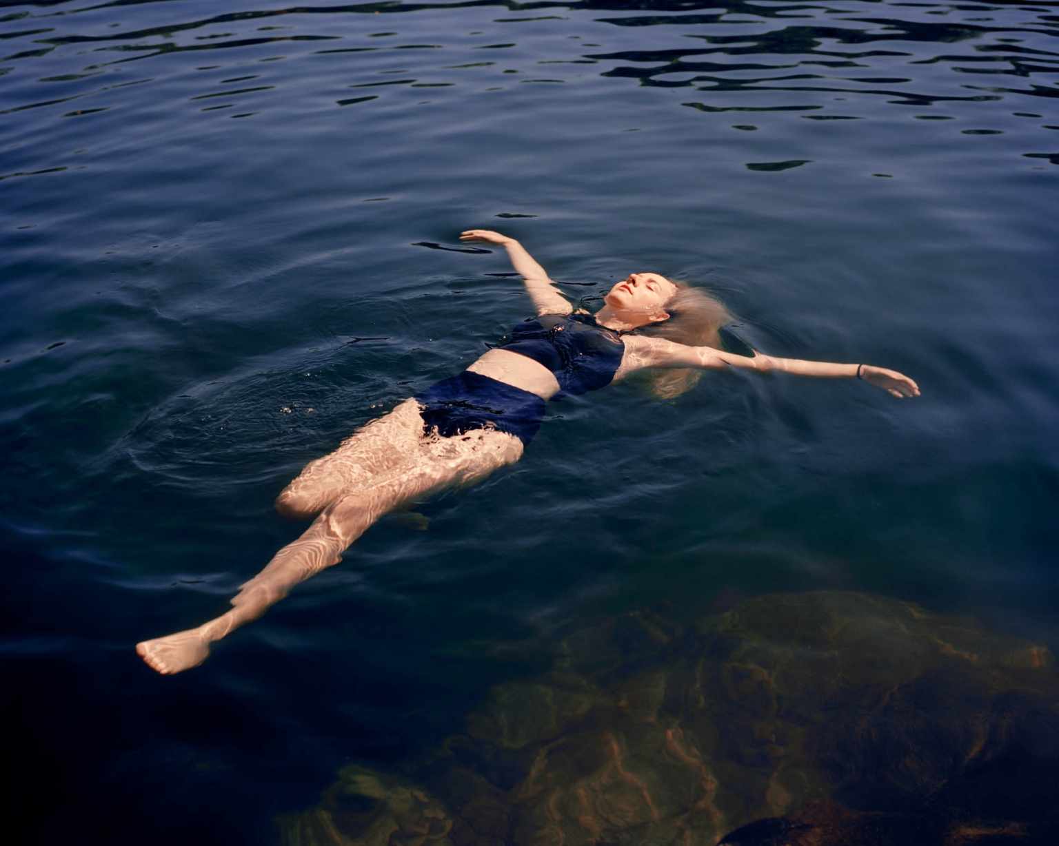 Adult woman floats on her back in dark blue water, arms outstretched. The setting is outdoors with natural light and visible rock formations.