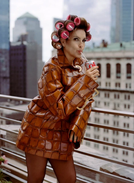 Woman with hair rollers drinking soda on a rooftop with a city skyline in the background wearing a brown patterned jacket.